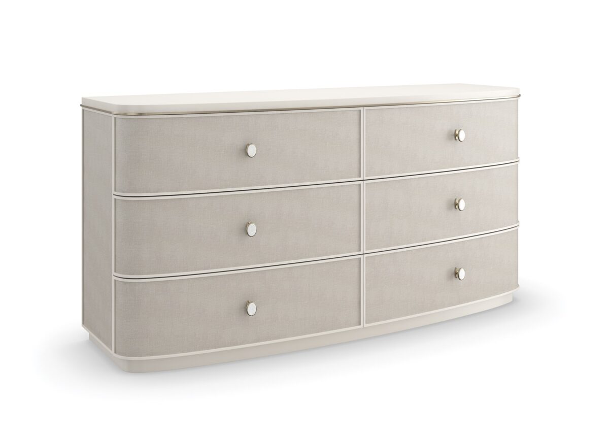 Lyon Dresser by Caracole — a 64″‑wide sculptural dresser in Almond Milk faux‑linen vinyl finish, with softly curved sloping sides and six vinyl‑wrapped drawers featuring painted Interiors, all set on a clean plinth base and trimmed with a slender Brushed‑Gold metal bead.