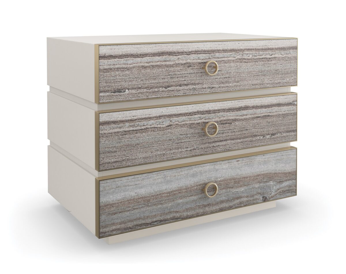 Bedrock Nightstand by Caracole — a 34″‑wide three‑drawer nightstand with polished gray sandstone fronts featuring subtle champagne‑gold trim and ring pulls, set in a creamy Almond Milk wood frame with a matching plinth base and soft‑close undermount drawer guides.