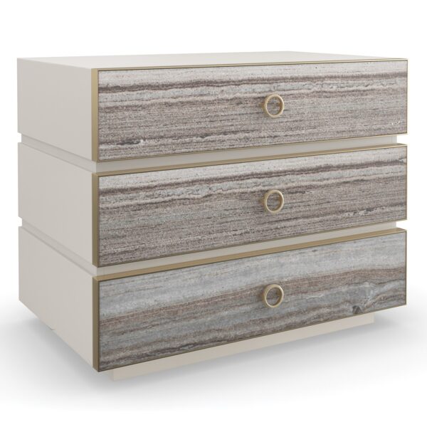 Bedrock Nightstand by Caracole — a 34″‑wide three‑drawer nightstand with polished gray sandstone fronts featuring subtle champagne‑gold trim and ring pulls, set in a creamy Almond Milk wood frame with a matching plinth base and soft‑close undermount drawer guides.
