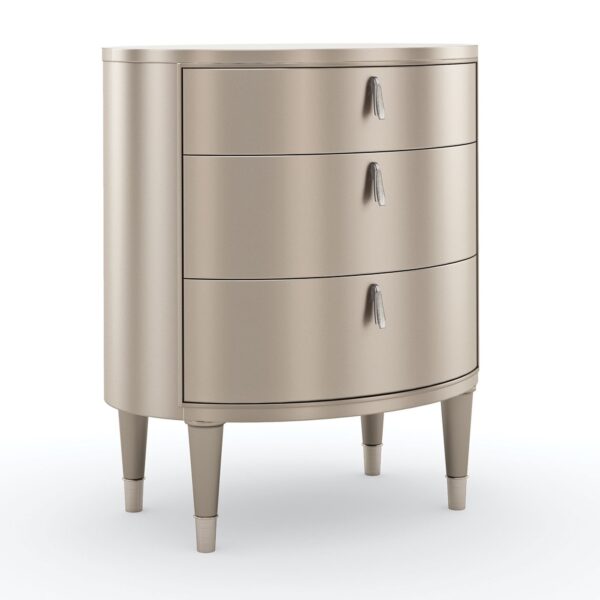 Bedside Beauty Nightstand by Caracole — a 24″-wide oval nightstand with three soft‑close drawers, finished in shimmering soft‑silver paint on Asian hardwood, topped with a softly curved top, accented by polished stainless‑steel ferrules on tapered legs, and elongated Art‑Deco pendant-style drawer pulls with faux-stone centers.