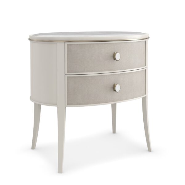 Bruges Nightstand by Caracole — a 28″‑wide oval bedside chest featuring a beveled Guangxi marble top, two vinyl‑wrapped drawers with linen-like texture and brushed‑gold metal bead inlay, set in an Almond Milk painted acacia wood case with jewelry-style round pulls, soft‑close undermount guides, and Almond Milk–painted interiors.