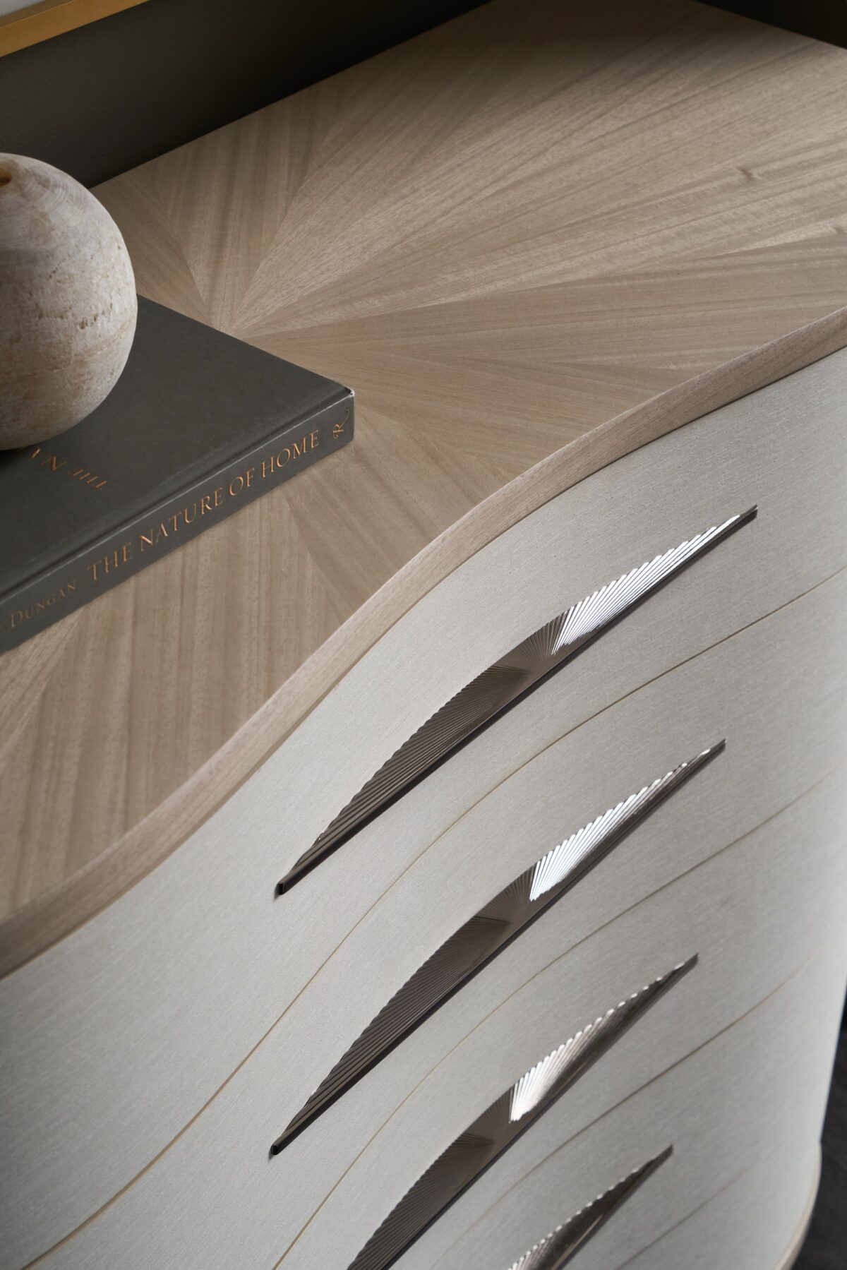 Meandrous Dresser by Caracole — a 50″‑wide organically curved dresser with five vinyl‑wrapped drawers in pearlescent Moonstone finish, set in a subtle Seal Skin‑tone plinth base; features horizontal bronze-tone handles, soft‑close undermount guides, painted interiors, a radial‑pattern Koto veneer top, and a removable jewel tray in the top drawer.