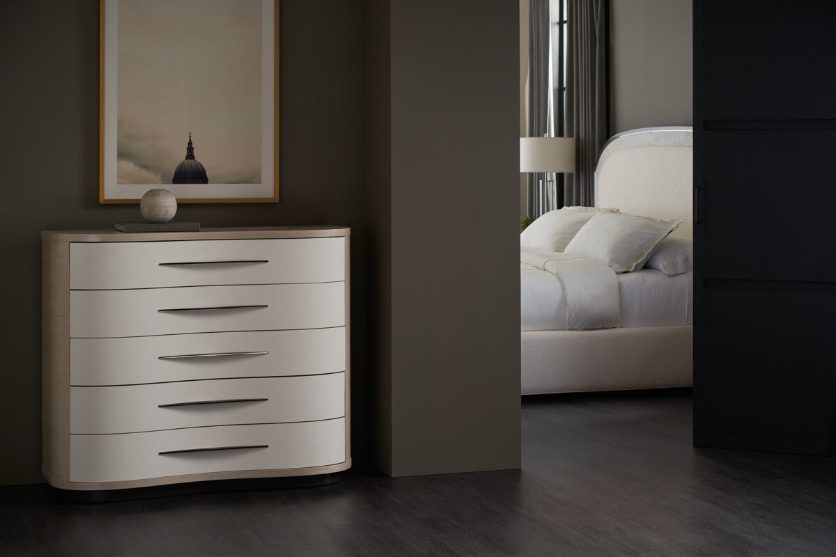 Meandrous Dresser by Caracole — a 50″‑wide organically curved dresser with five vinyl‑wrapped drawers in pearlescent Moonstone finish, set in a subtle Seal Skin‑tone plinth base; features horizontal bronze-tone handles, soft‑close undermount guides, painted interiors, a radial‑pattern Koto veneer top, and a removable jewel tray in the top drawer.