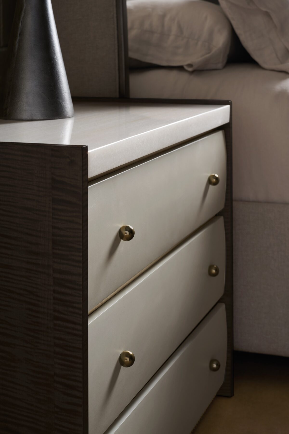 Avignon Bronze Nightstand by Caracole — a 34″‑wide classic nightstand with three soft‑close drawers featuring tactile Rockport Burnished Adobe fronts, set within Craftsman Bronze‑painted end panels; topped with striated white Guangxi stone, trimmed with antique‑bronze‑leaf dentil molding at the base, brushed‑gold ball knobs, and painted Almond Milk interiors.