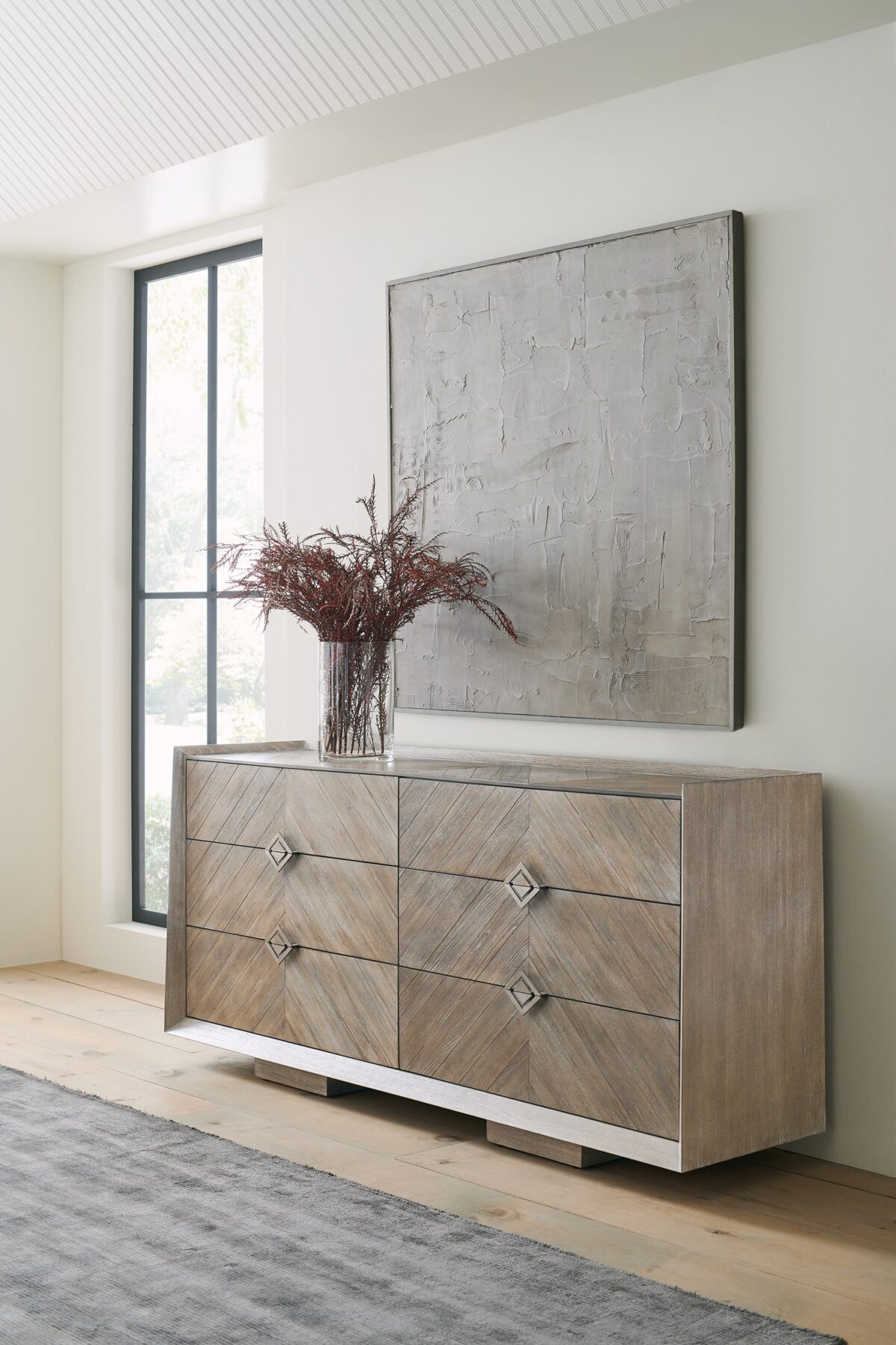 Naturally Dresser by Caracole — a 74″‑wide six‑drawer dresser in Ash Driftwood finish with chevron‑patterned wood‑veneer fronts, accented by a slender Sundance Gold metal bead edging and recessed drawer pulls, all sitting on low block feet.