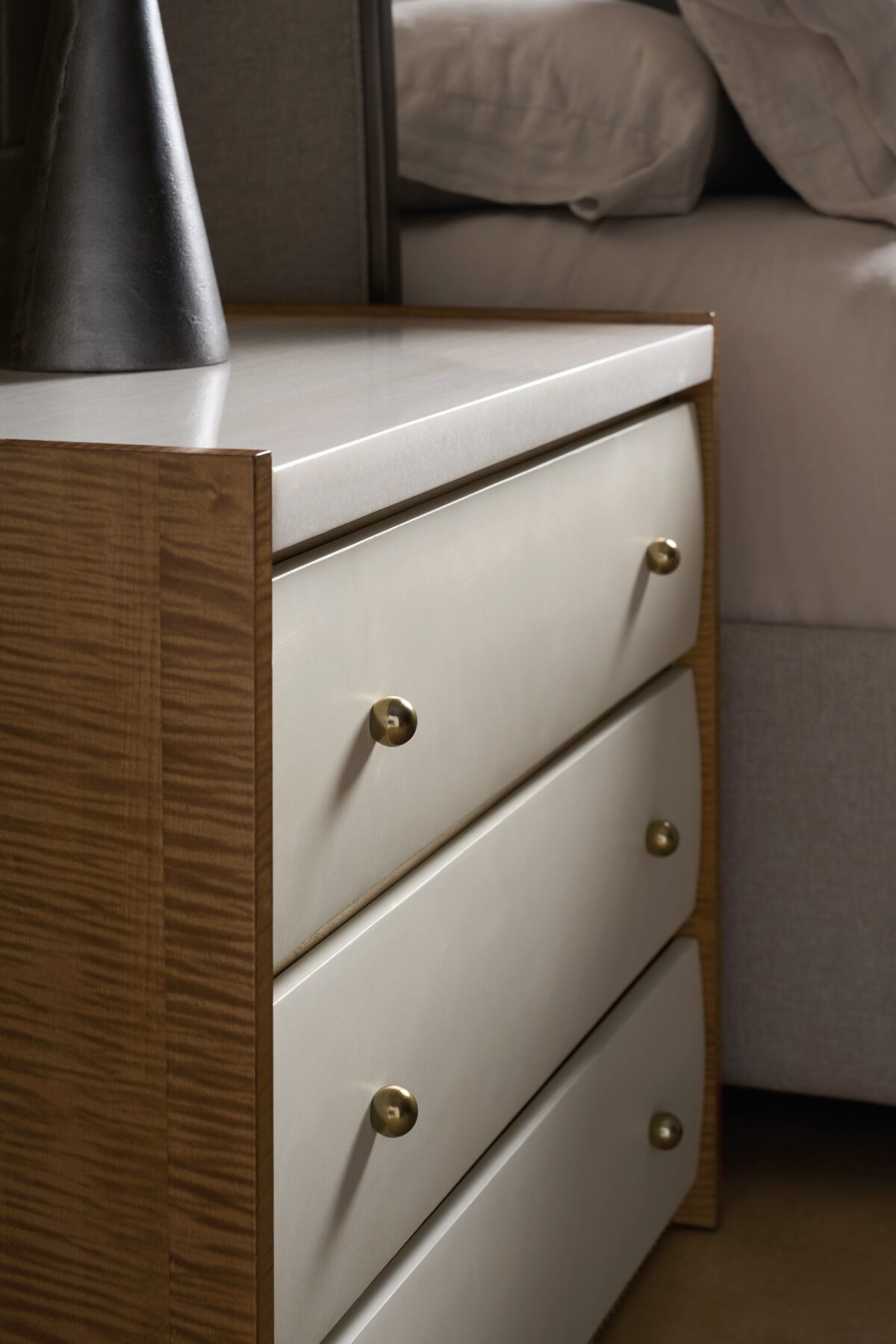 Caracole Avignon Nightstand — a 34″‑wide three‑drawer nightstand with tactile Rockport Burnished Adobe drawer fronts, topped with a striated white Guangxi/onyx stone surface, flanked by Toasted Sycamore end panels, adorned with antique‑bronze‑leaf dentil molding at the base, and brushed‑gold ball knobs on soft‑close undermount guides.