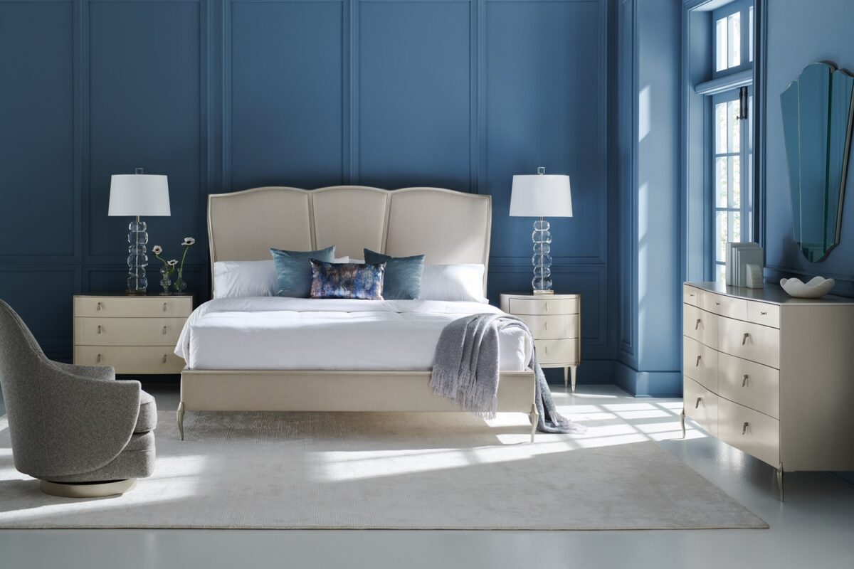 Bedside Beauty Nightstand by Caracole — a 24″-wide oval nightstand with three soft‑close drawers, finished in shimmering soft‑silver paint on Asian hardwood, topped with a softly curved top, accented by polished stainless‑steel ferrules on tapered legs, and elongated Art‑Deco pendant-style drawer pulls with faux-stone centers.