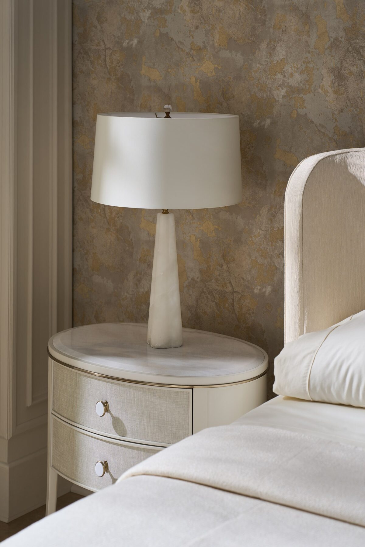 Bruges Nightstand by Caracole — a 28″‑wide oval bedside chest featuring a beveled Guangxi marble top, two vinyl‑wrapped drawers with linen-like texture and brushed‑gold metal bead inlay, set in an Almond Milk painted acacia wood case with jewelry-style round pulls, soft‑close undermount guides, and Almond Milk–painted interiors.