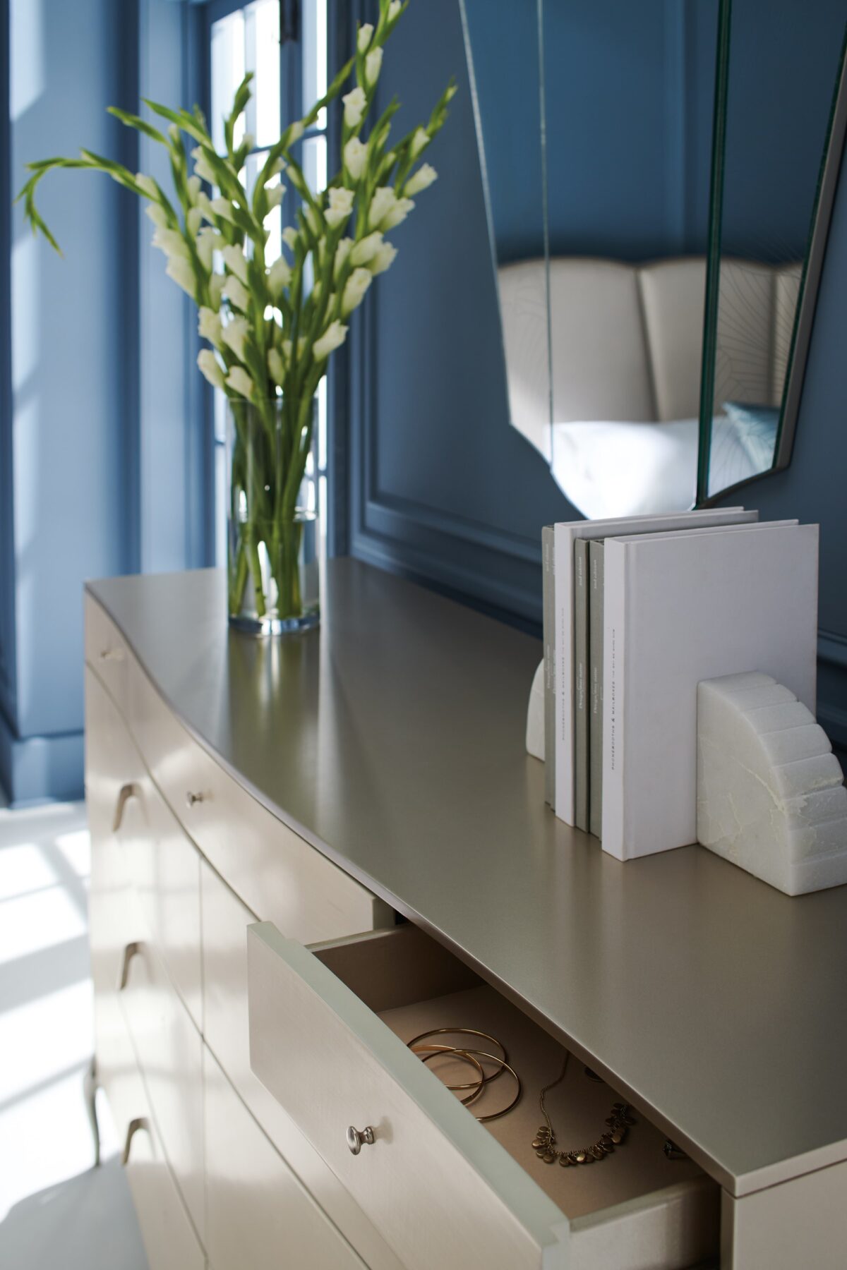 Dress to Impress Dresser by Caracole — a 62″ wide nine‑drawer dresser in soft‑silver painted Asian hardwood case, with vanilla‑cream figured‑sycamore drawer fronts, elongated Art‑Deco style metal pulls, a gently curved top, ornate silver‑toned legs, and two felt‑lined top drawers.