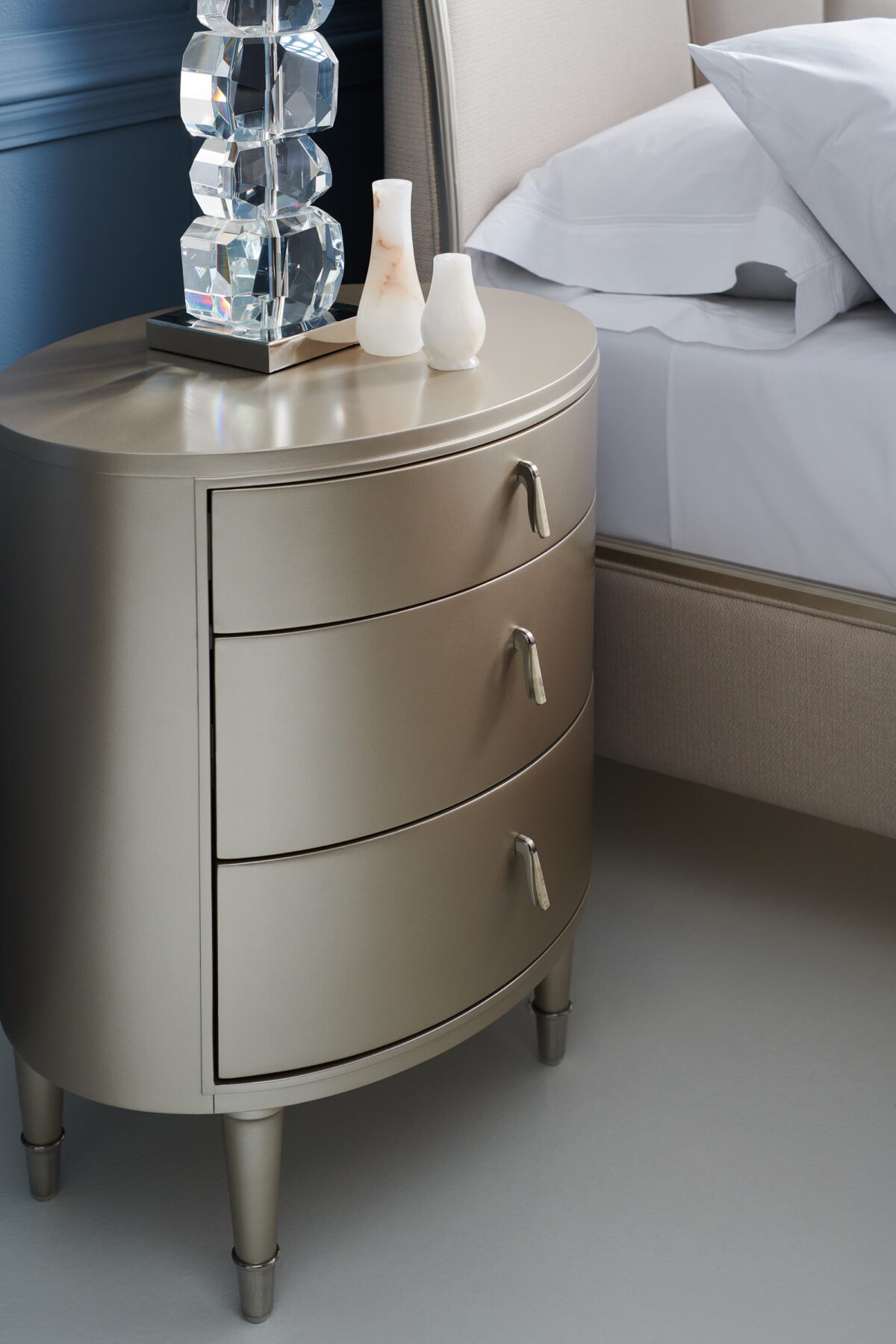 Bedside Beauty Nightstand by Caracole — a 24″-wide oval nightstand with three soft‑close drawers, finished in shimmering soft‑silver paint on Asian hardwood, topped with a softly curved top, accented by polished stainless‑steel ferrules on tapered legs, and elongated Art‑Deco pendant-style drawer pulls with faux-stone centers.