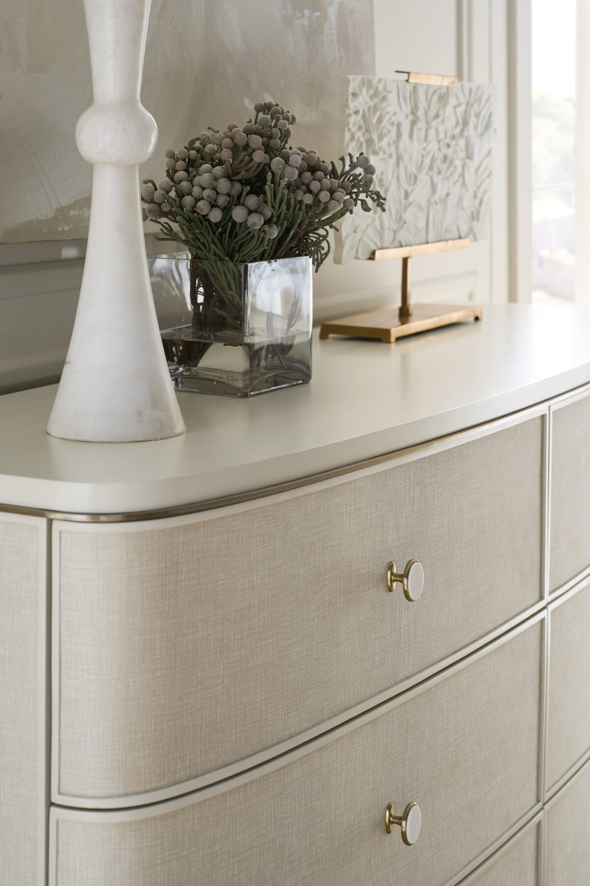 Lyon Dresser by Caracole — a 64″‑wide sculptural dresser in Almond Milk faux‑linen vinyl finish, with softly curved sloping sides and six vinyl‑wrapped drawers featuring painted Interiors, all set on a clean plinth base and trimmed with a slender Brushed‑Gold metal bead.
