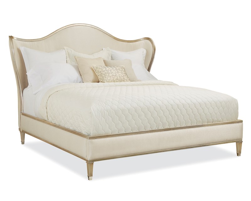 Caracole Bedtime Beauty King Bed featuring a camel-back headboard with curving wings, fully upholstered in cream strie fabric, and an exposed wood rail finished in Auric silver leaf.