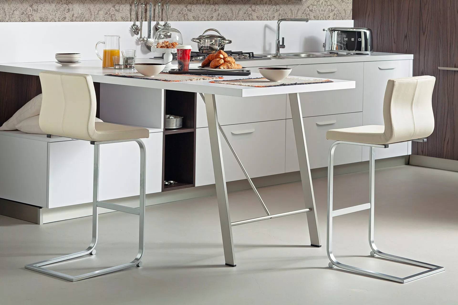 Ocean bar stool by Peressini upholstered in warm grey fabric with a curved low back and brushed stainless steel pedestal base, staged at a white quartz kitchen island.