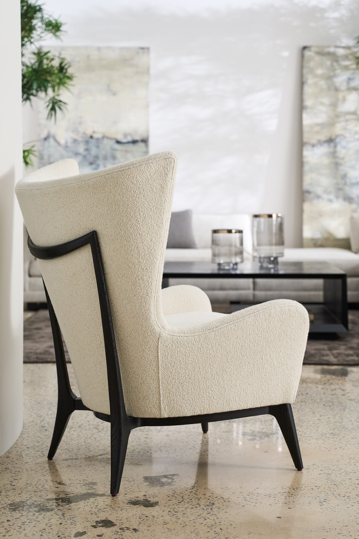 The Caracole "Whats New Pussycat?" wing chair, featuring a modern Scandinavian design with a light-colored upholstered body, gracefully sloped arms, a soft round back, and a distinctive exposed wood detail on the outback in a Modern Walnut or Almost Black finish.