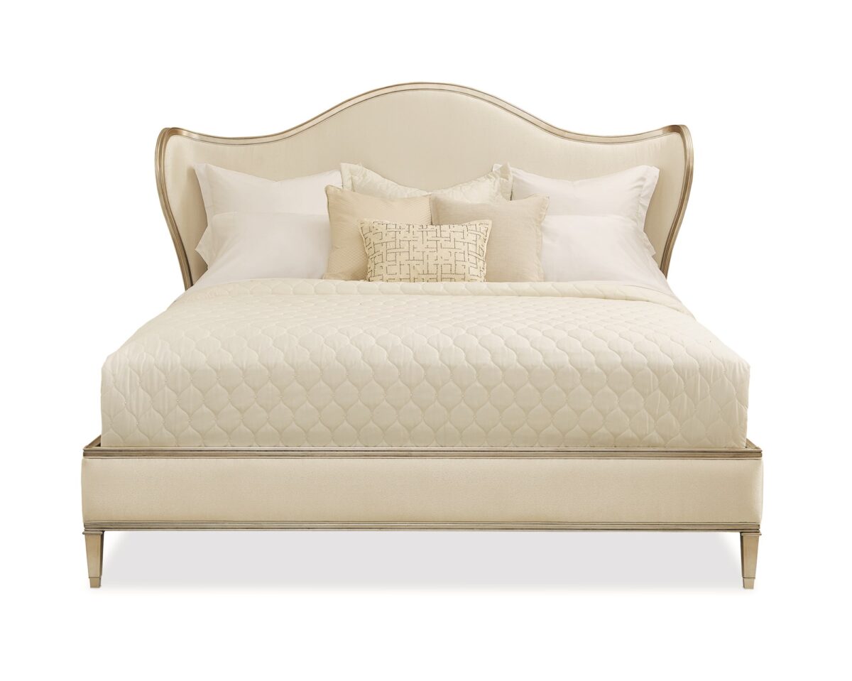 Caracole Bedtime Beauty King Bed featuring a camel-back headboard with curving wings, fully upholstered in cream strie fabric, and an exposed wood rail finished in Auric silver leaf.