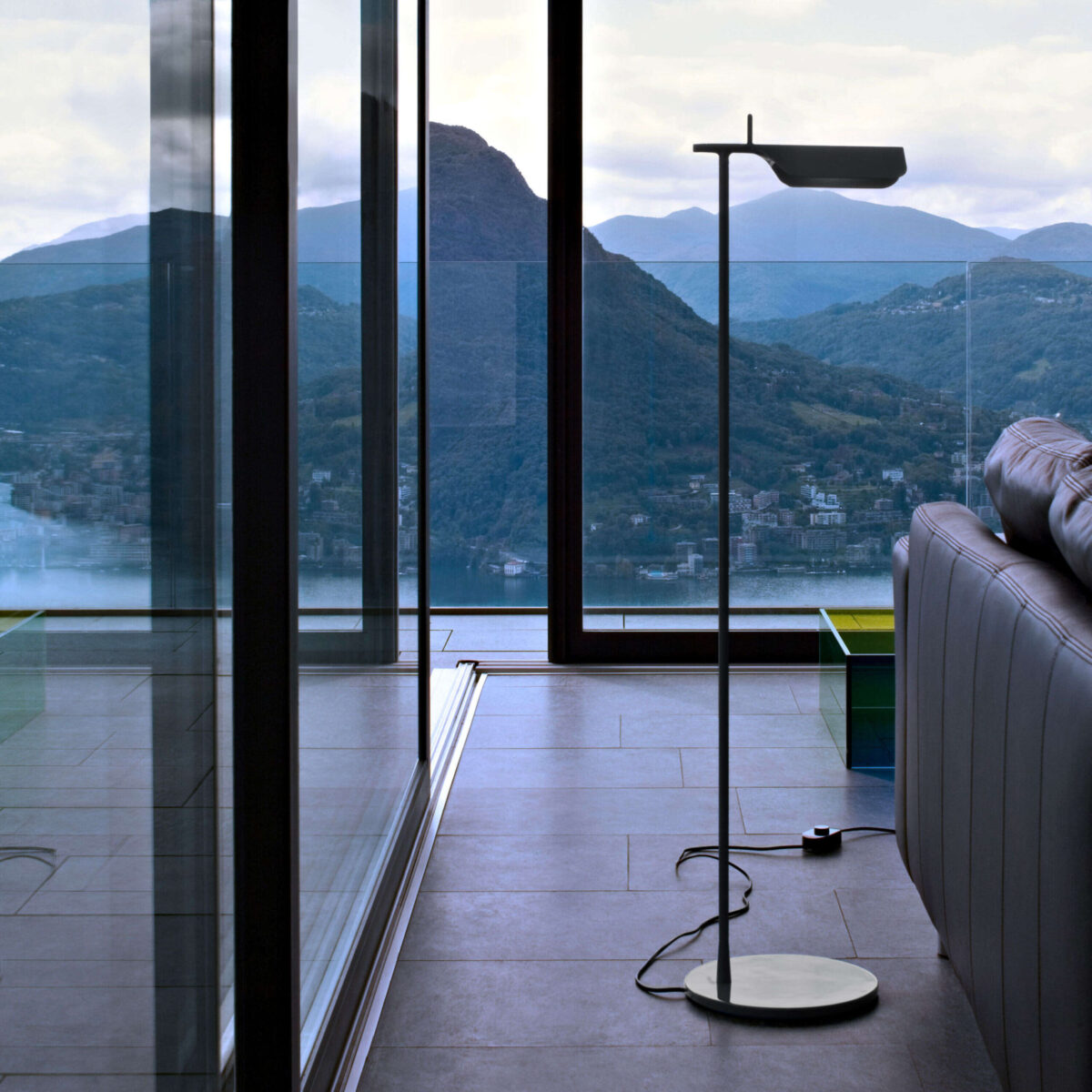 Black Tab Floor lamp positioned beside a sofa, the pivoting slim head casting focused direct light over a reading nook.