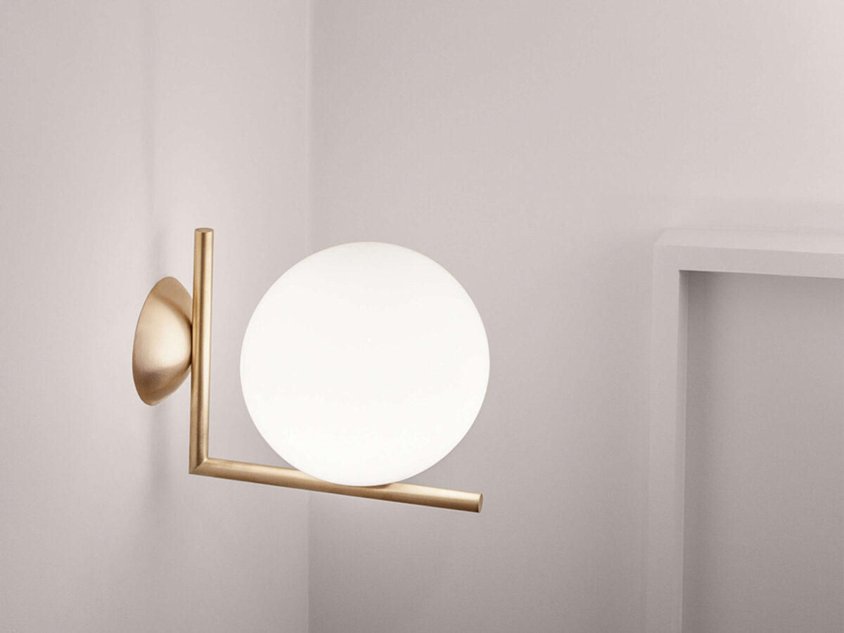 A Flos IC Lights C/W2 lamp, showcasing its distinctive design with a perfectly spherical white glass shade resting on a brushed brass L-shaped arm, creating a sophisticated and balanced aesthetic.